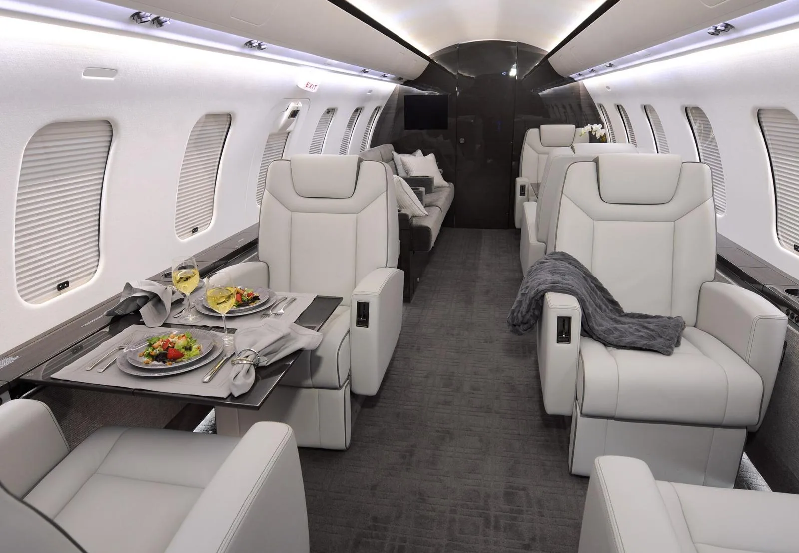 The luxurious interior of a private jet cabin features light grey leather seats, a dining table set with food and drinks, and a dark grey carpet.