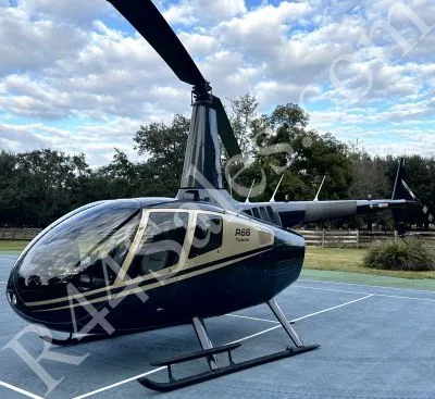 A dark blue and gold Robinson R66 Turbine helicopter is parked on a paved surface with trees in the background.