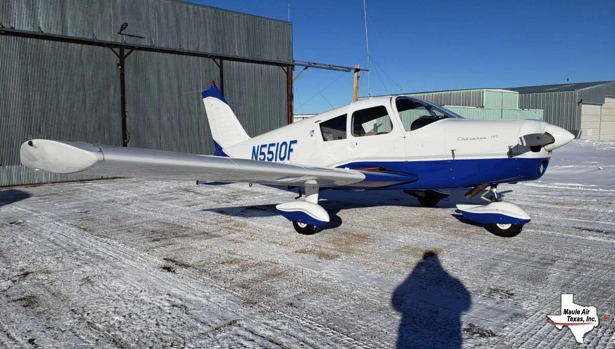 A white and blue Piper Cherokee 140 aircraft (N551OF) parked on a snowy tarmac in front of a hangar.