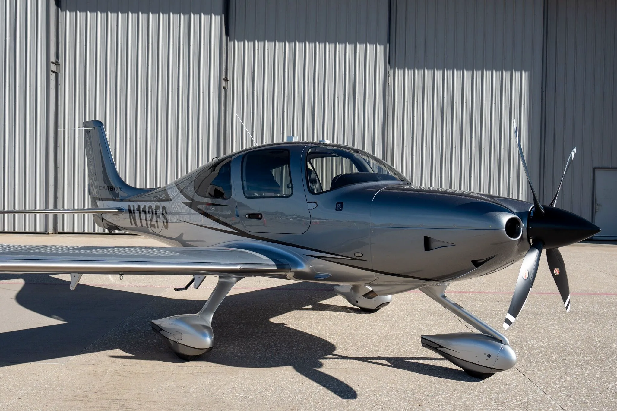 A front-right view of a silver and dark grey Cirrus SR22T Carbon Edition aircraft (N112FS) with a hangar in the background.