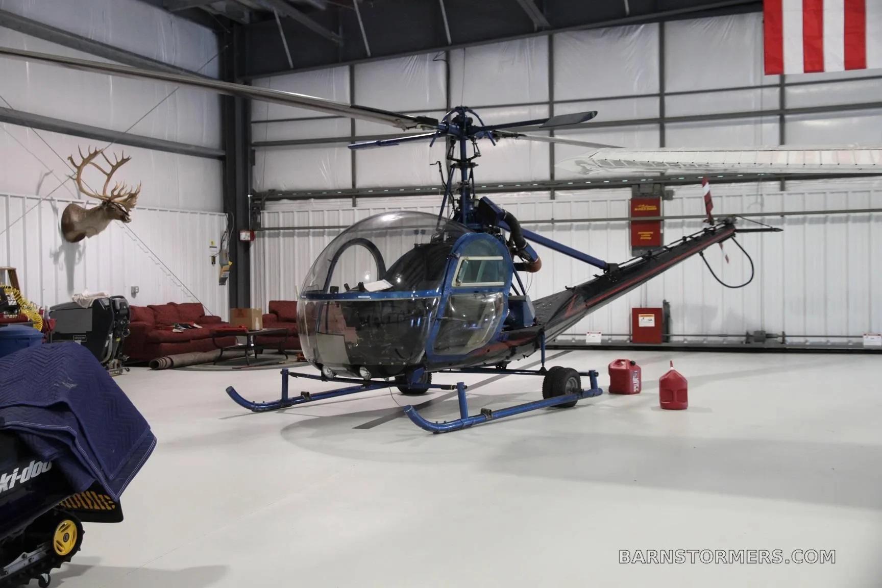 A blue and black helicopter with an open cockpit sits inside a hangar, with a deer head mount on the wall.