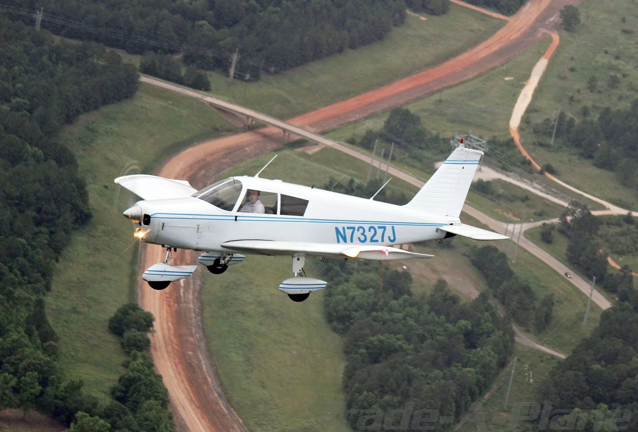 A white Piper Cherokee 140 aircraft, N7327J, with blue stripes, flies over a landscape with roads and trees.