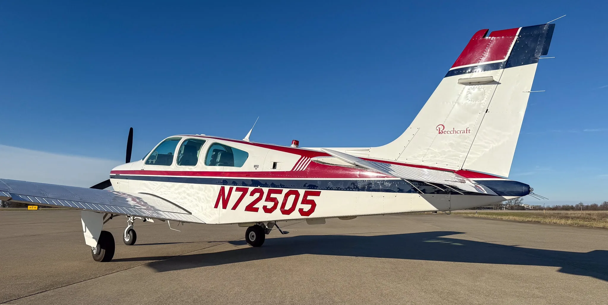 Rear-right view of a white Beechcraft Bonanza with red and blue stripes, showing registration N72505 and Beechcraft logo on the tail.