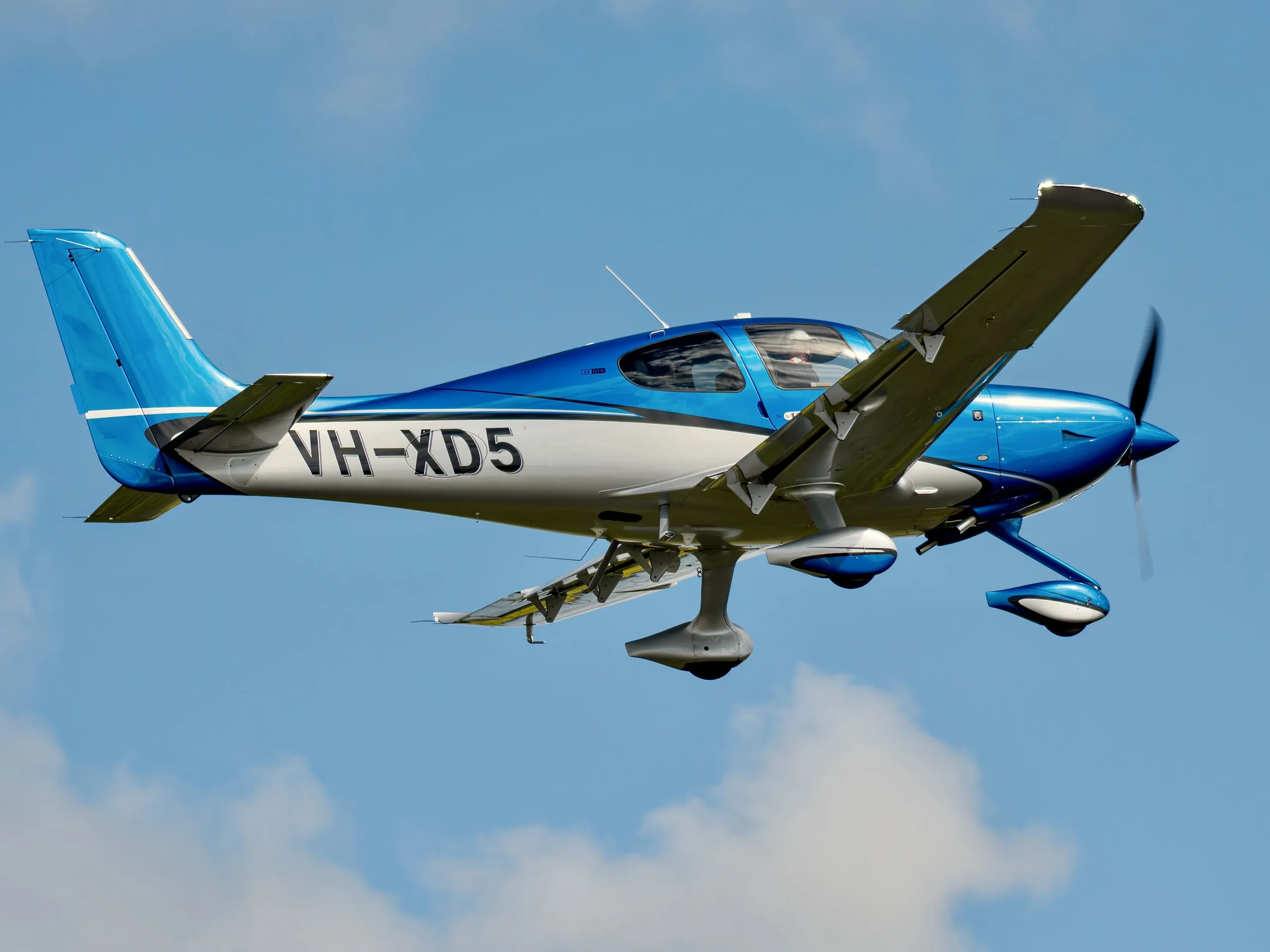 A blue and white single-engine Cirrus SR22 aircraft with registration VH-XD5 flying against a blue sky with clouds.
