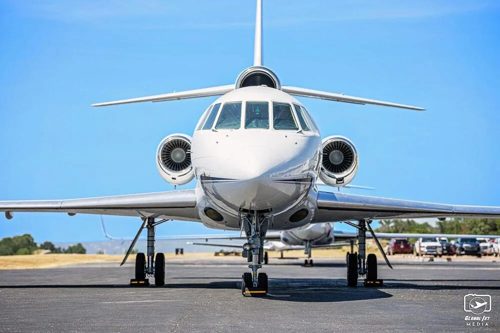 A head-on view of a white Dassault Falcon 900EX business jet with blue stripes, parked on an airfield.