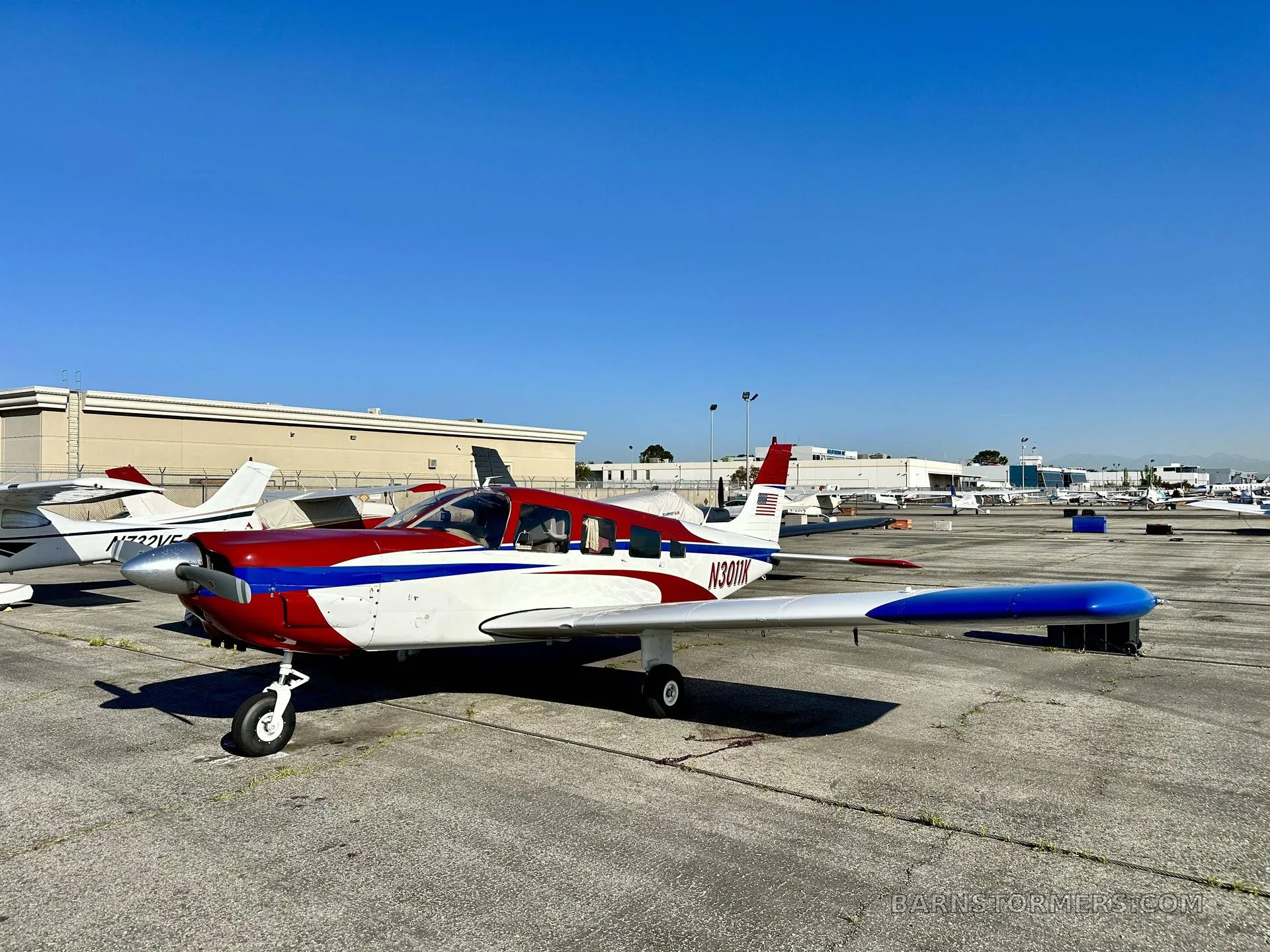 A red, white, and blue single-engine propeller aircraft (N3011K) parked on an airport ramp under a clear blue sky.