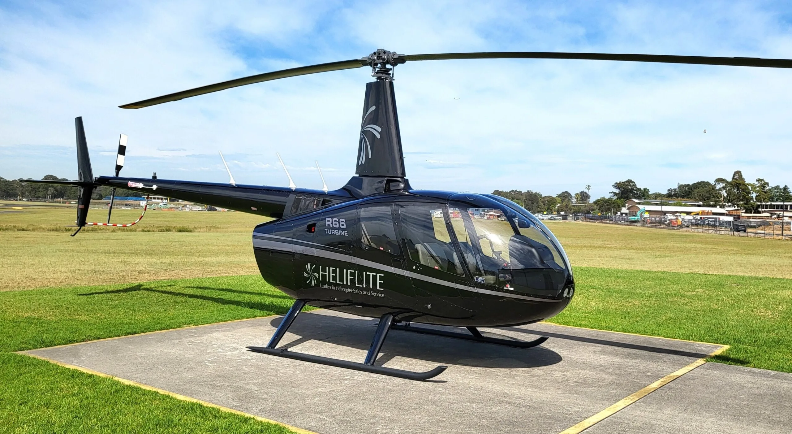 A black Robinson R66 Turbine helicopter with "HELIFLITE" branding is parked on a concrete pad at an airfield.