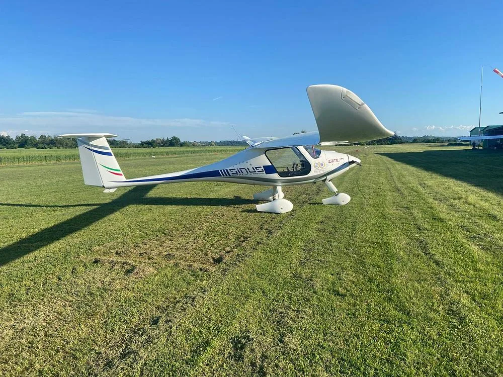 A white Pipistrel Sinus aircraft with blue and green tail stripes, parked on a sunny green airfield.