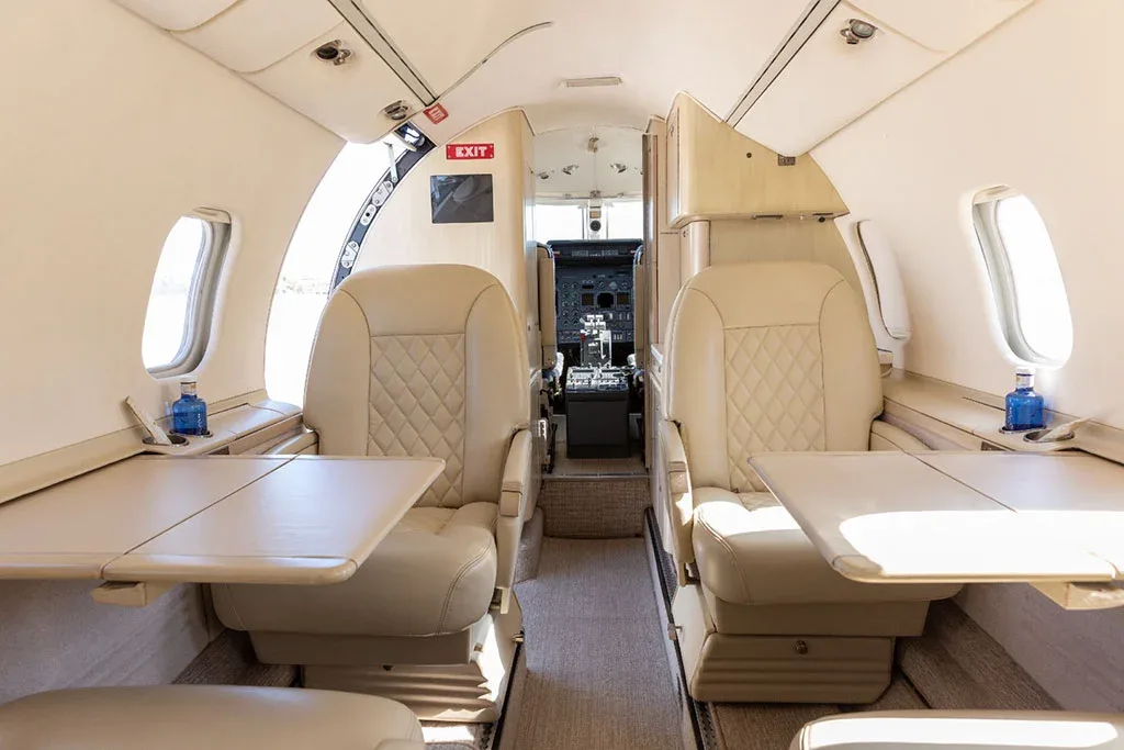 The elegant cabin interior of a Learjet, showing beige quilted leather seats, fold-out tables, and a view into the cockpit.