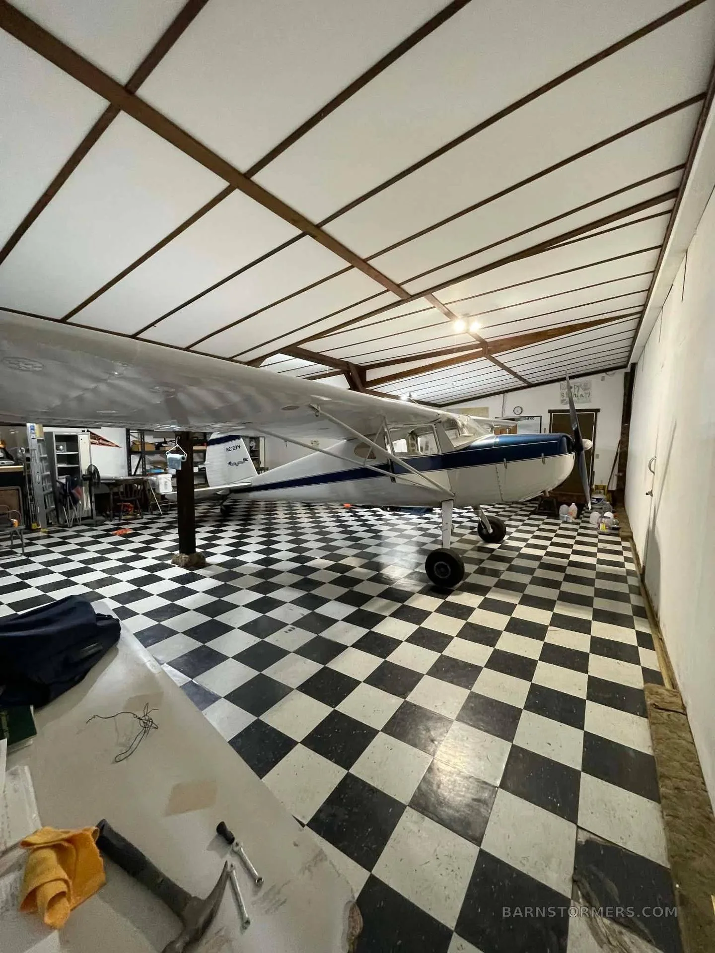 A white and blue taildragger aircraft is parked inside a hangar with a black and white checkered floor.