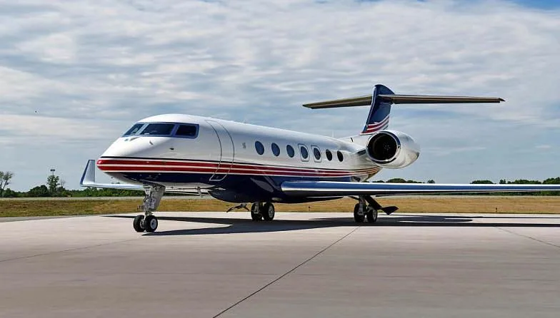 A white private jet with blue and red stripes is parked on a tarmac under a partly cloudy sky.