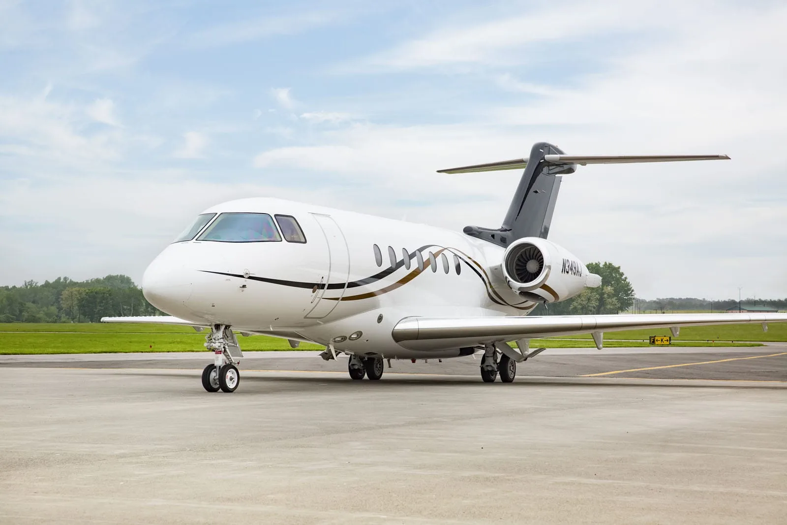 A white business jet with dark grey and gold accents on the tail and fuselage, parked on an asphalt runway under a partly cloudy sky.