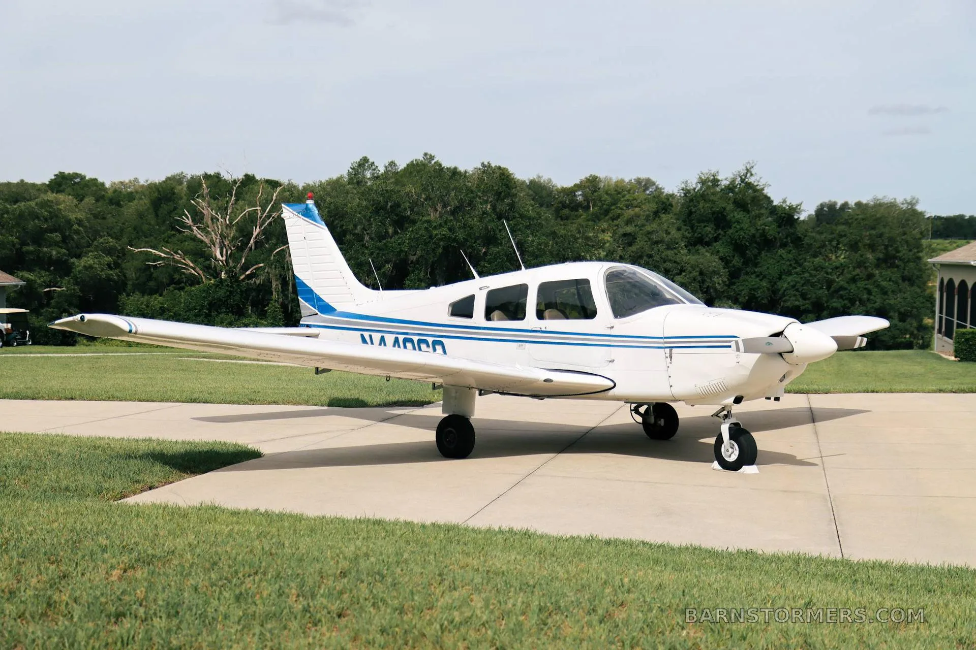 A white Piper PA-28 series aircraft with blue stripes, parked on a concrete pad with trees in the background.