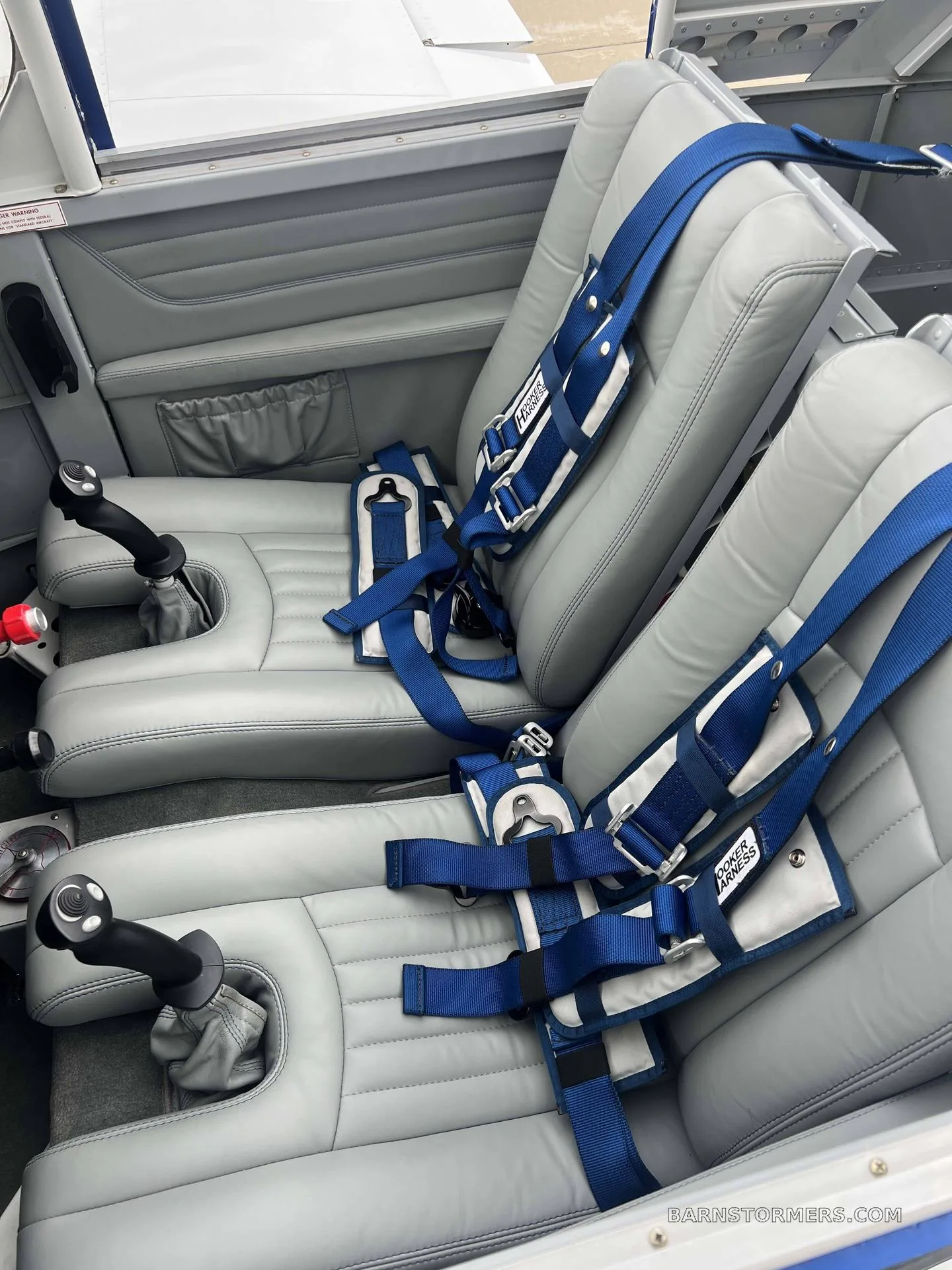 The interior cabin of a light aircraft, showing two grey leather seats with blue "HOOKER HARNESS" seatbelts and dual stick controls.