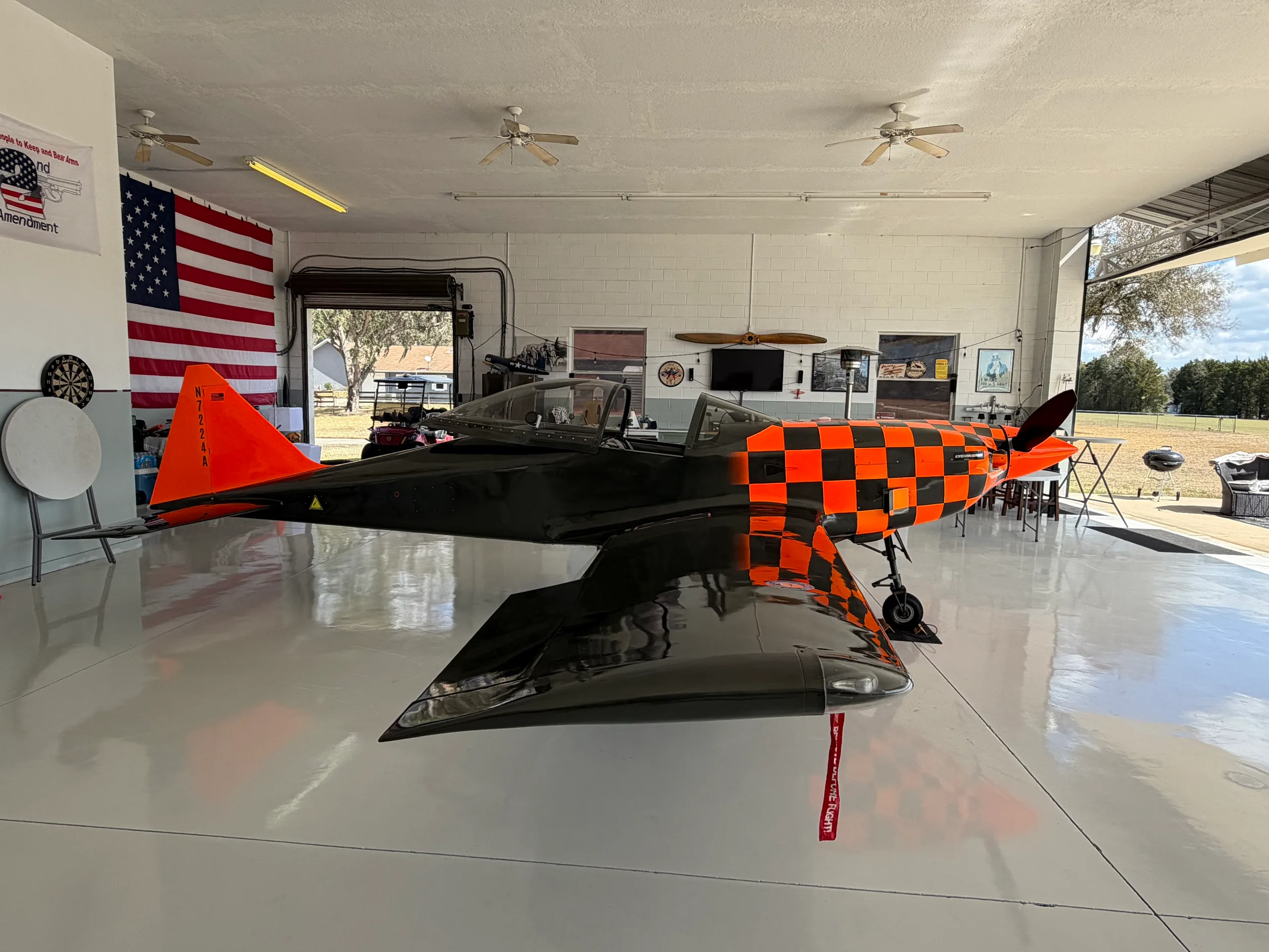 A full side view of the black and orange checkered experimental aircraft, N7224A, parked inside a hangar with an American flag.