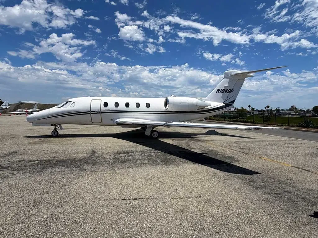 A white Cessna Citation business jet (N184GP) with a dark stripe, parked on an asphalt tarmac under a blue sky with scattered clouds.