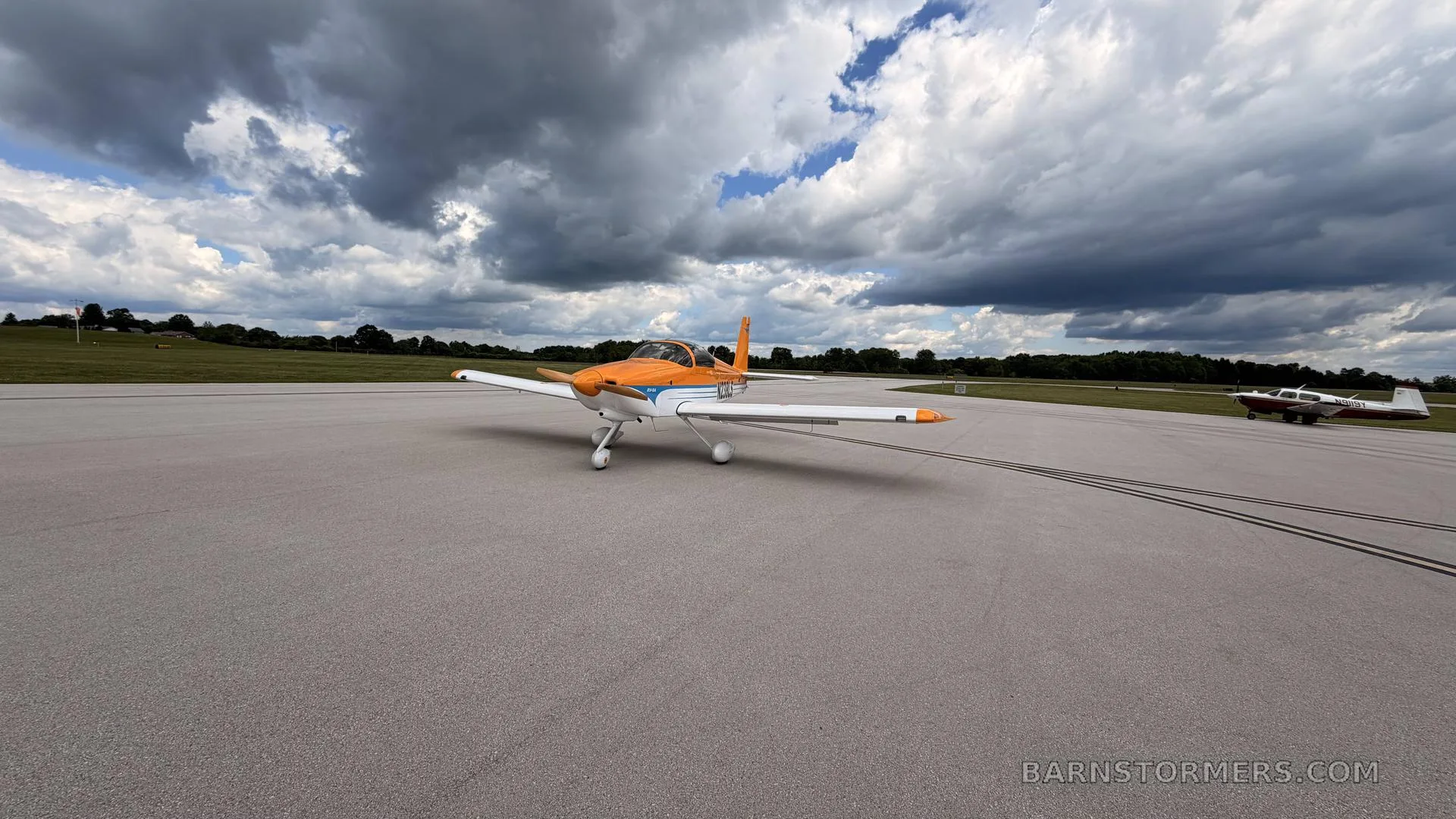 A low-wing, single-engine aircraft with an orange and white paint scheme on an airfield under a cloudy sky, with another aircraft in the background.