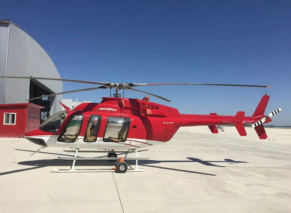 A red and white Bell 407GXP helicopter is parked on a tarmac with a hangar in the background under a clear blue sky.
