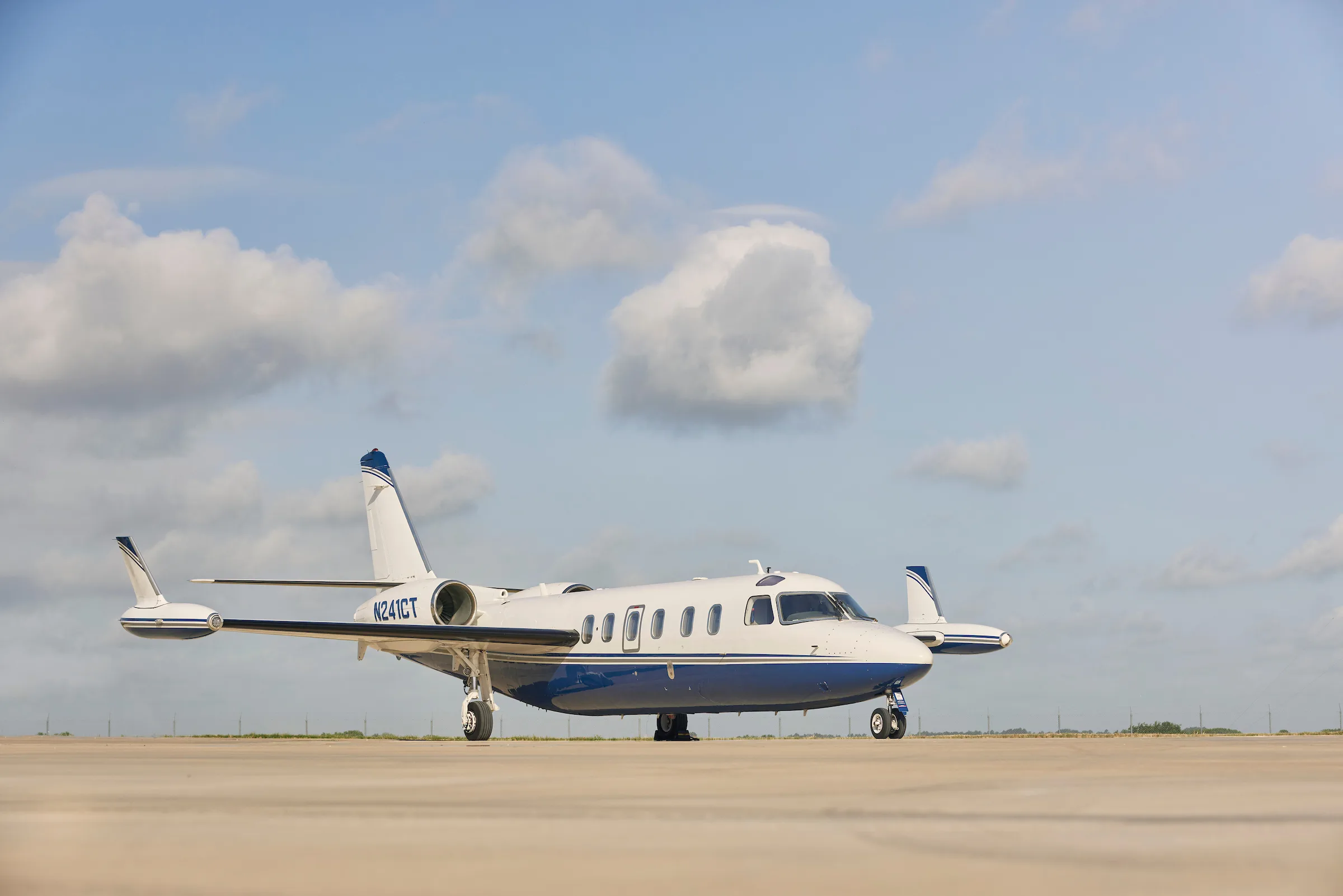 A white and blue business jet, tail number N241CT, is parked on an airfield under a partly cloudy sky.