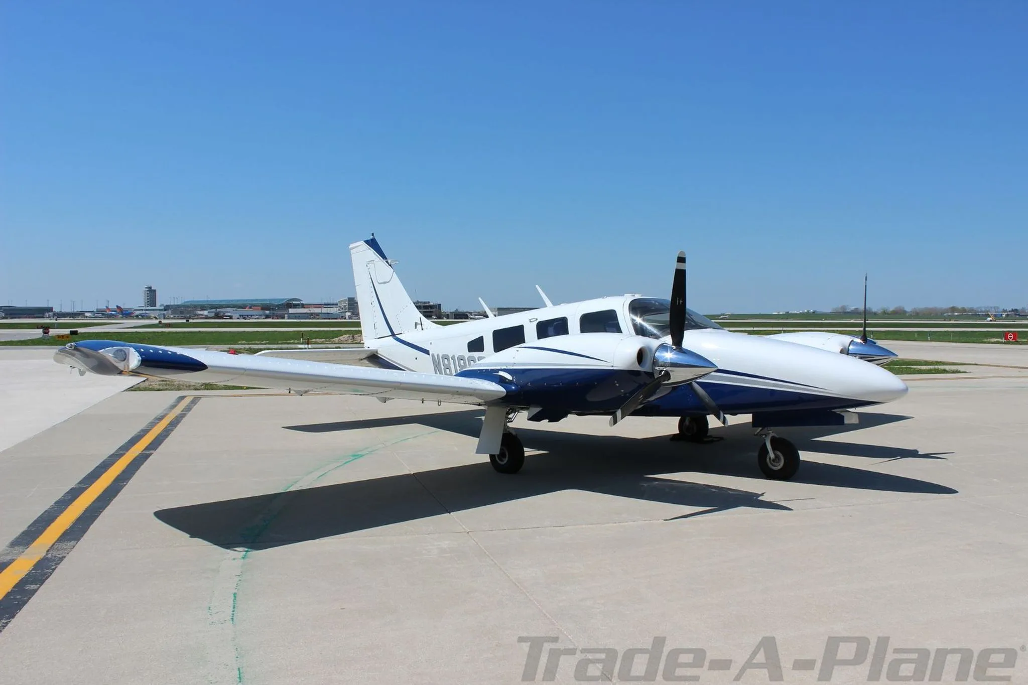 A white and blue twin-engine Piper Seneca aircraft, registration N8186R, parked on an airport tarmac under a clear blue sky.