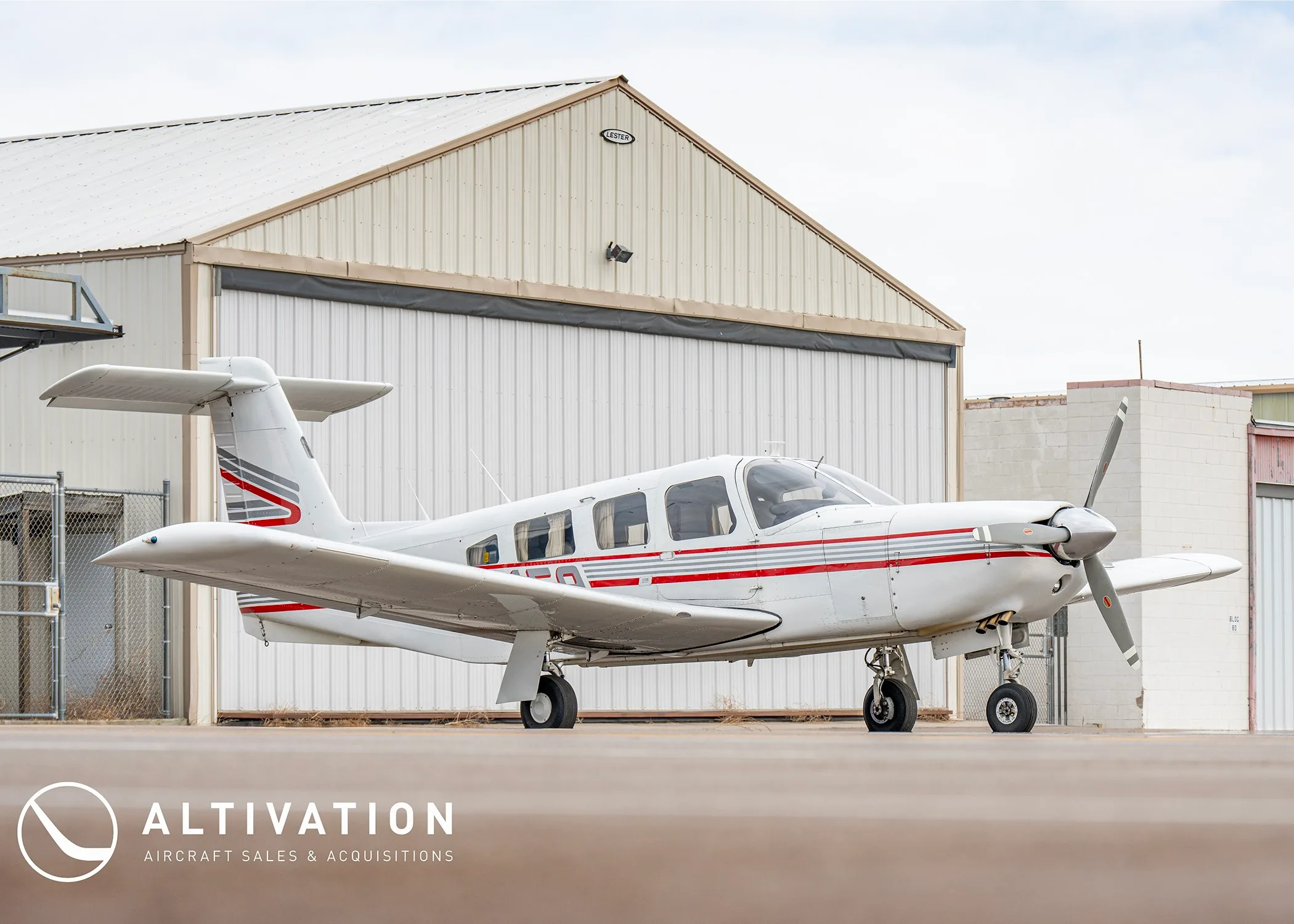 A white single-engine propeller aircraft with red and grey stripes parked on an apron next to a hangar.