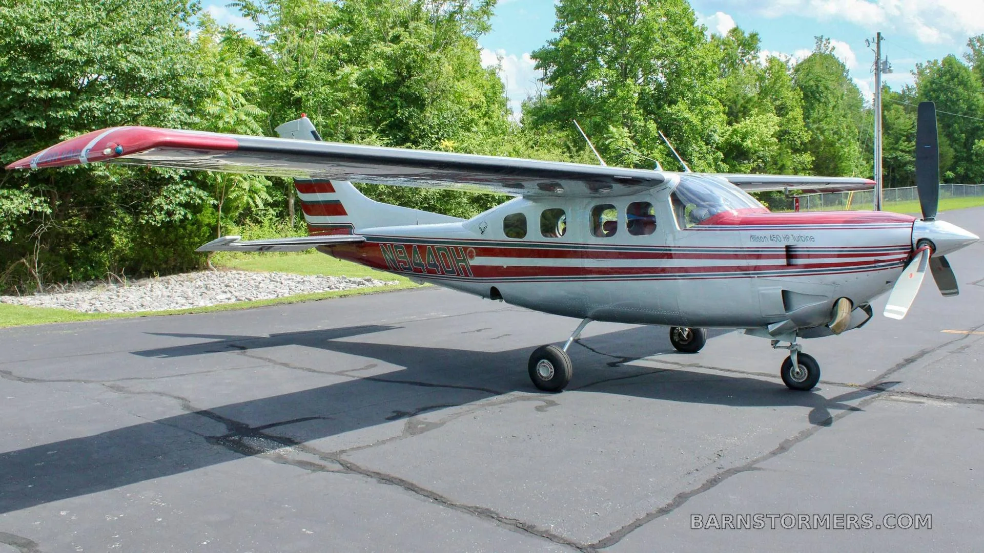 A silver and red single-engine high-wing aircraft with registration N944DH is parked on an asphalt ramp with trees in the background.