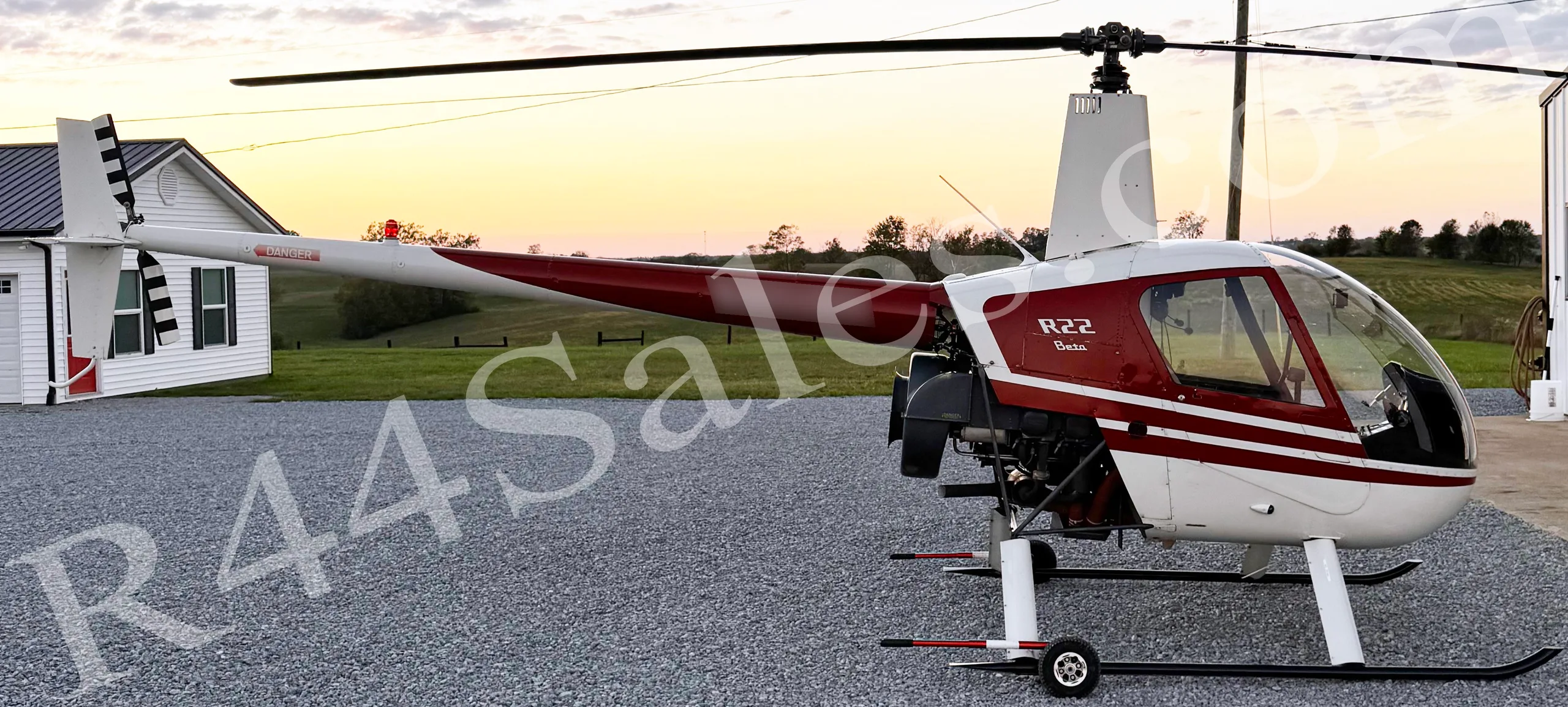 A white and red Robinson R22 Beta helicopter parked on gravel at sunset with a white building in the background.