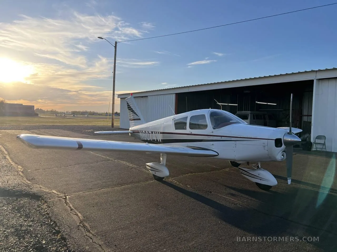 A white single-engine propeller aircraft with red and black stripes, parked outside a hangar at sunset.