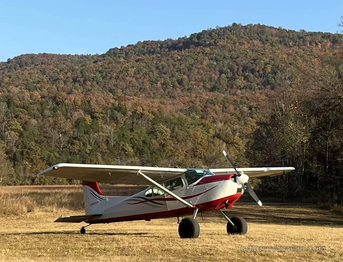 A white and red Cessna 180 aircraft with large tundra tires parked on a grassy field with autumn-colored mountains in the background.