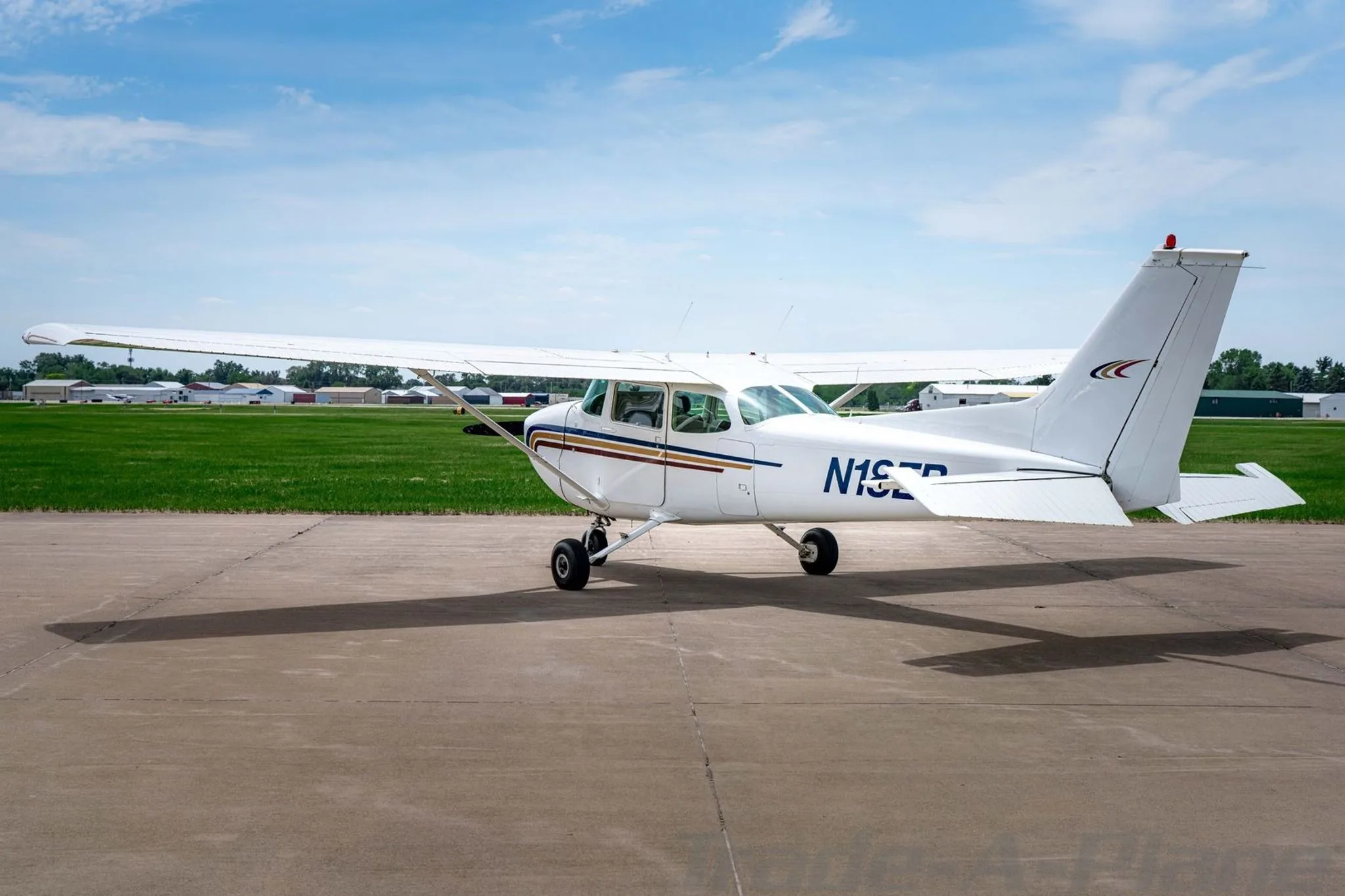 A white Cessna 172 with blue, gold, and red stripes on the fuselage, parked on an asphalt ramp, viewed from the rear-left.
