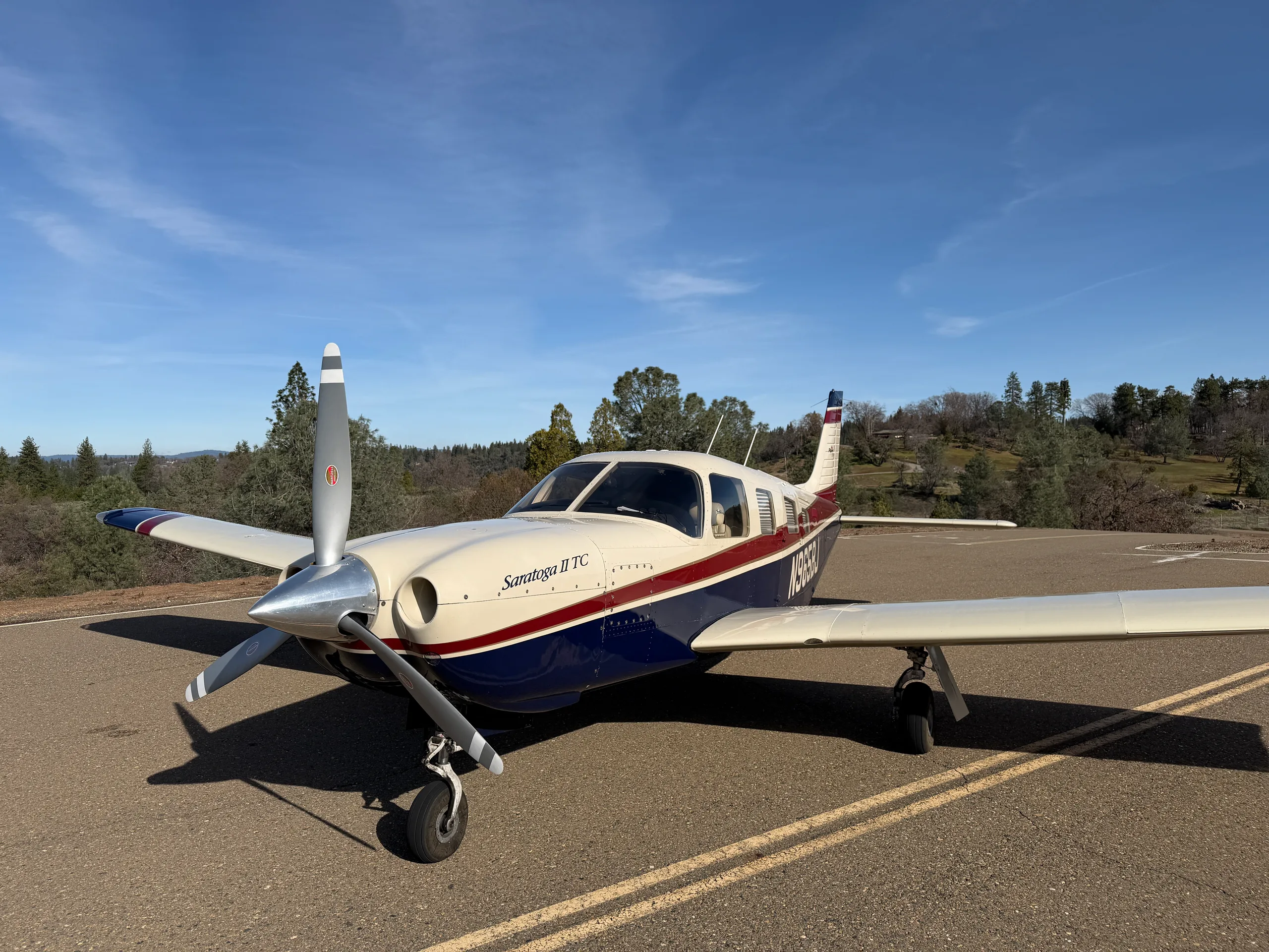 A Piper Saratoga II TC aircraft with a cream, blue, and red paint scheme parked on an asphalt surface under a clear blue sky.