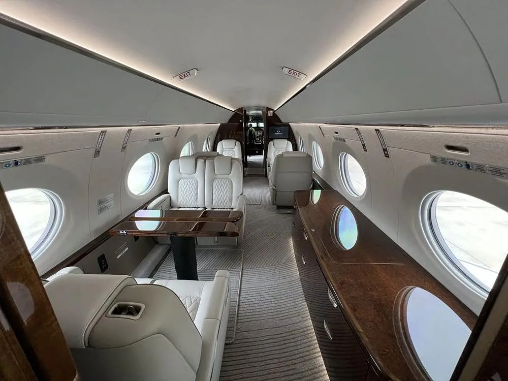 The luxurious interior of a private jet cabin with white quilted seats and dark wood tables.