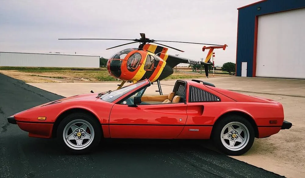 A red Ferrari sports car is parked in front of a Hughes 369HS helicopter with orange, yellow, and black stripes at an airfield.