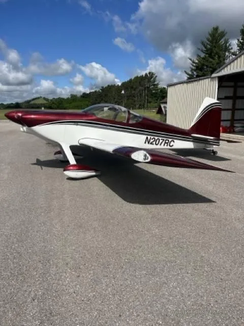 A side view of a red and white single-engine aircraft with tail number N207RC, parked on an asphalt apron next to a hangar.