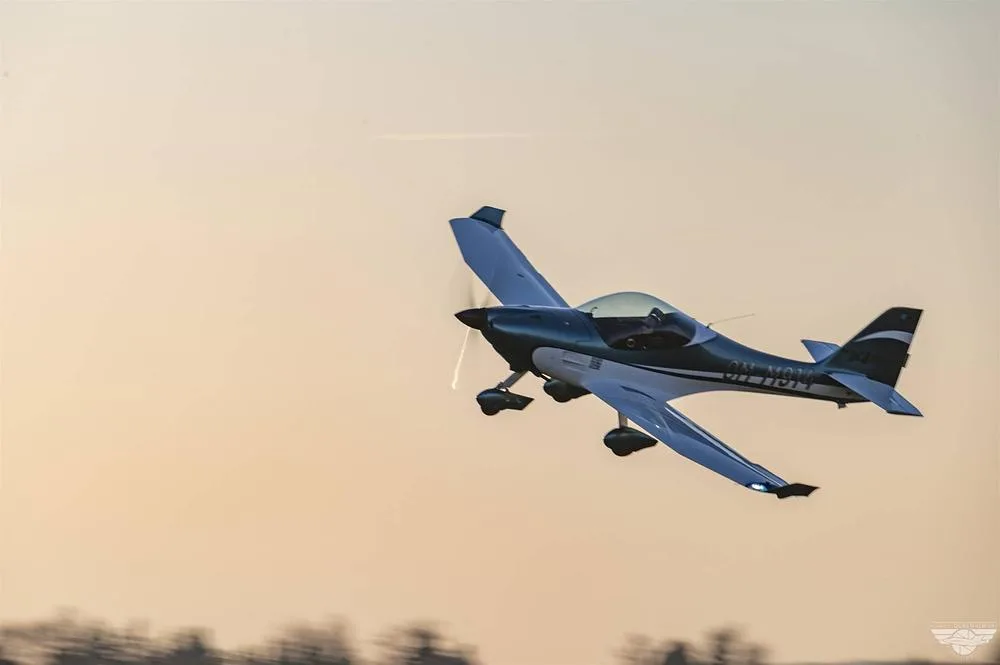 A blue and white FK14 Polaris aircraft (OM-M914) in flight, seen from below against a pale sky.