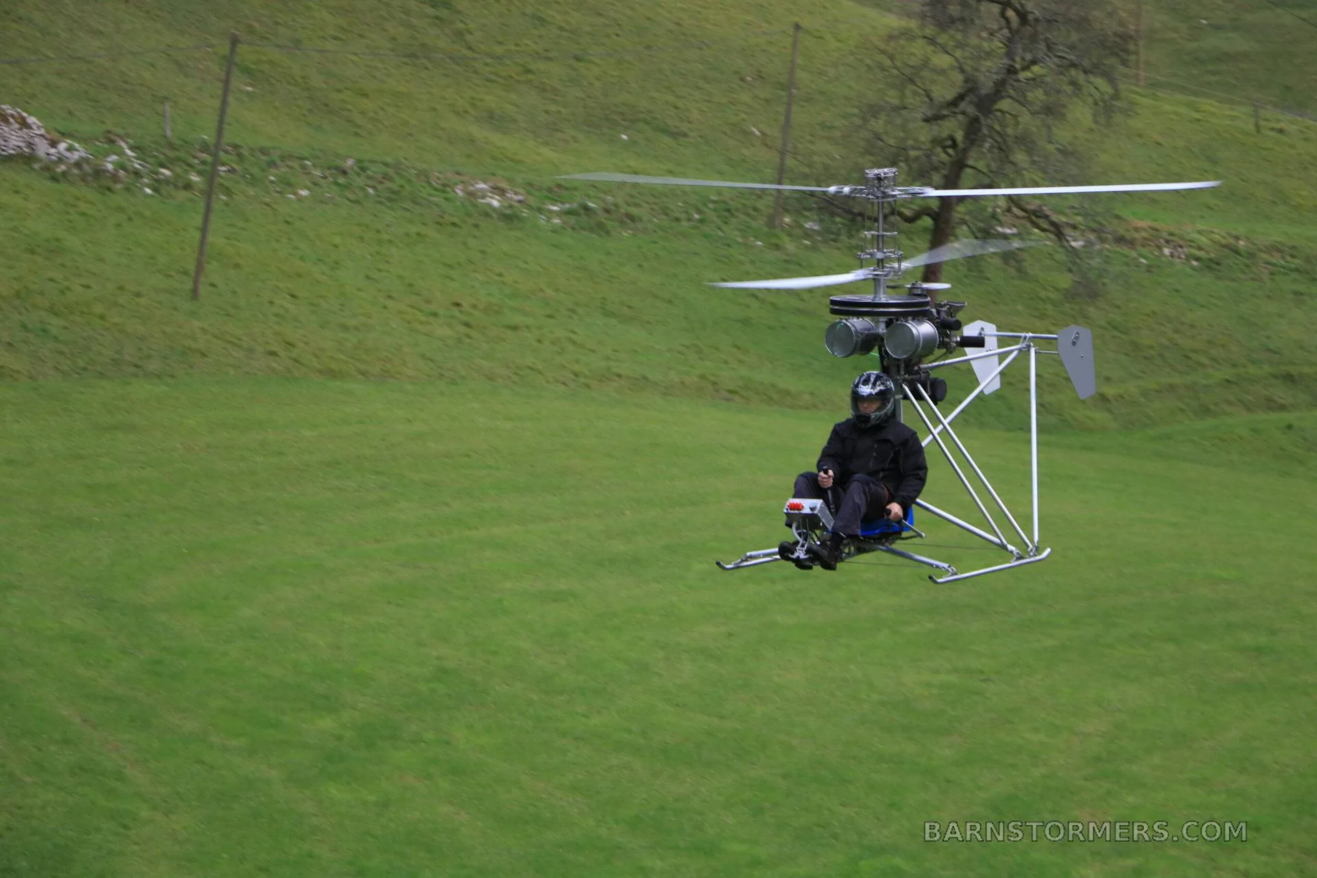 A single-seat ultralight helicopter with a pilot in black gear hovers over a green grassy field.