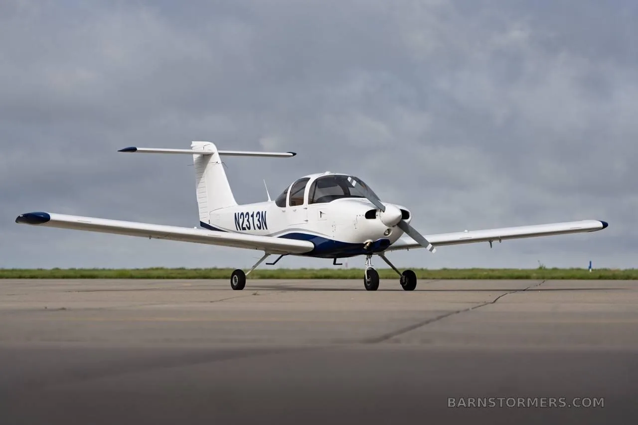 A white and blue single-engine propeller aircraft with registration N2313N is parked on an asphalt runway under a cloudy sky, viewed from the front-left.