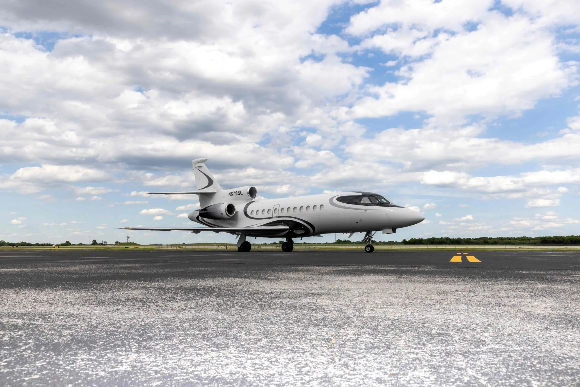 A white Dassault Falcon 900 private jet with black and grey accents is parked on an asphalt runway under a partly cloudy sky.