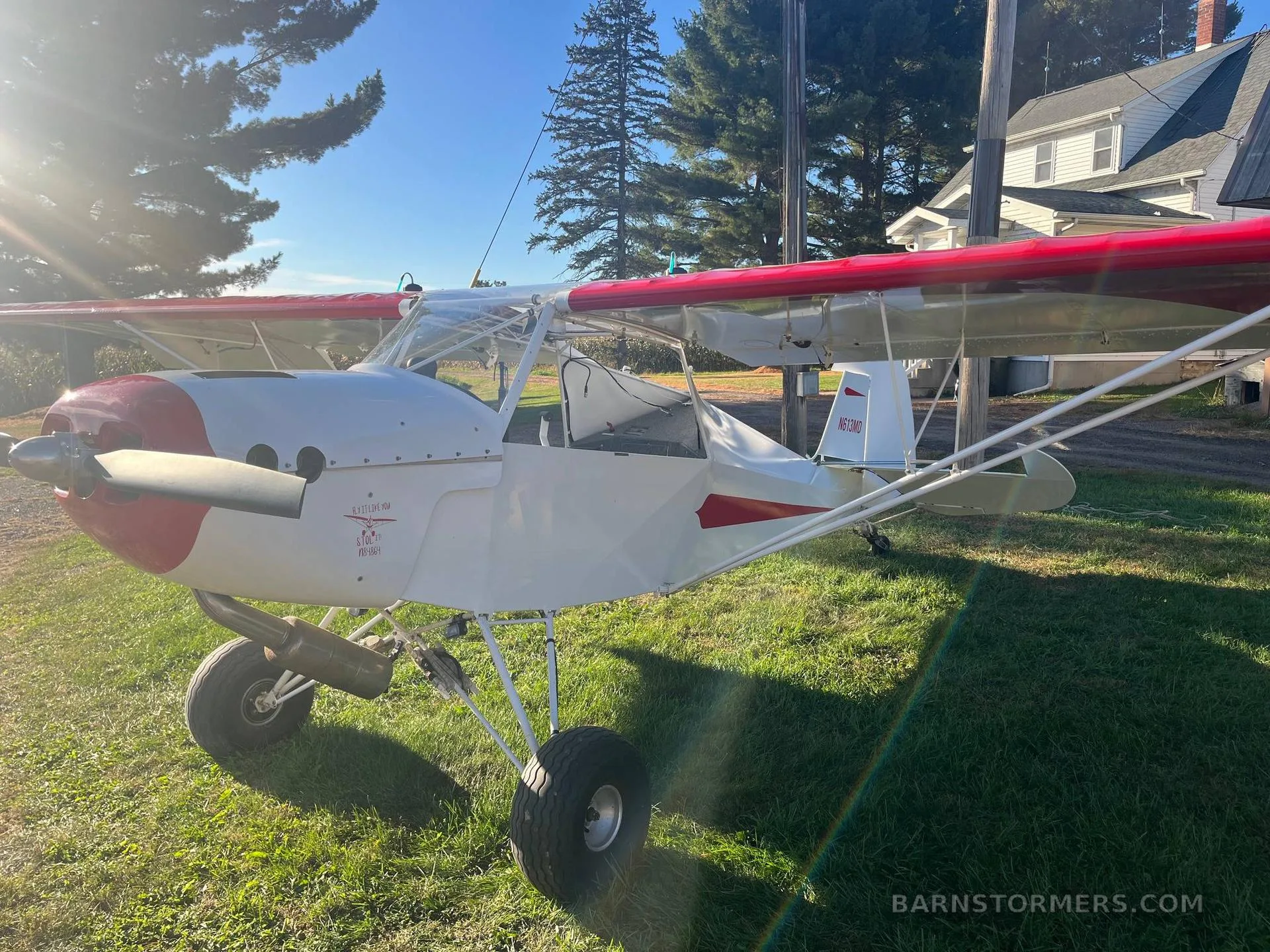 A white and red single-engine, high-wing experimental aircraft with large tires, parked on grass with a house in the background.