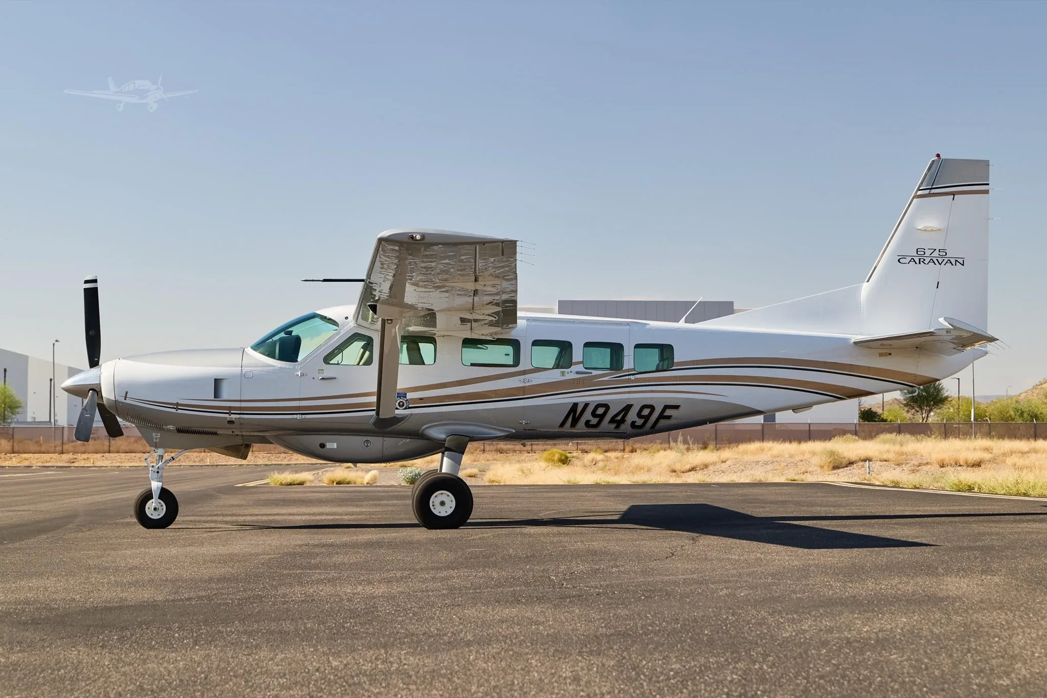 A white and gold Cessna 675 Caravan aircraft, registration N949F, is parked on an asphalt tarmac under a clear sky.