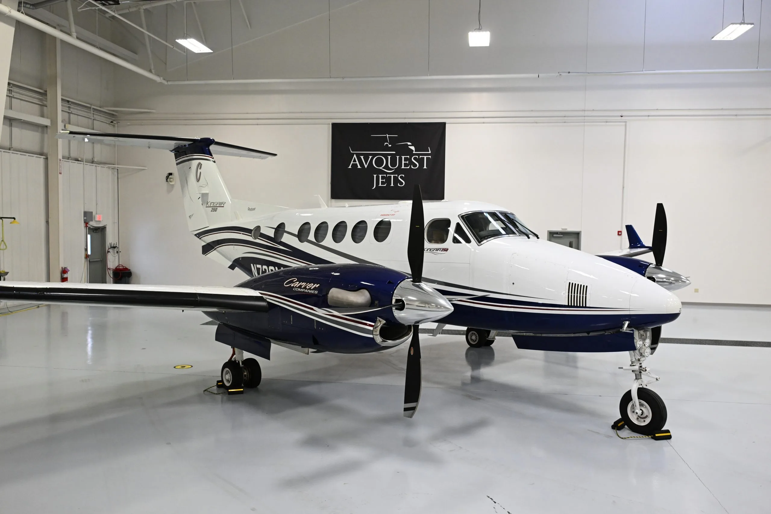 A white and blue Beechcraft King Air 250 turboprop aircraft with dark blue and grey stripes, parked inside a brightly lit hangar.
