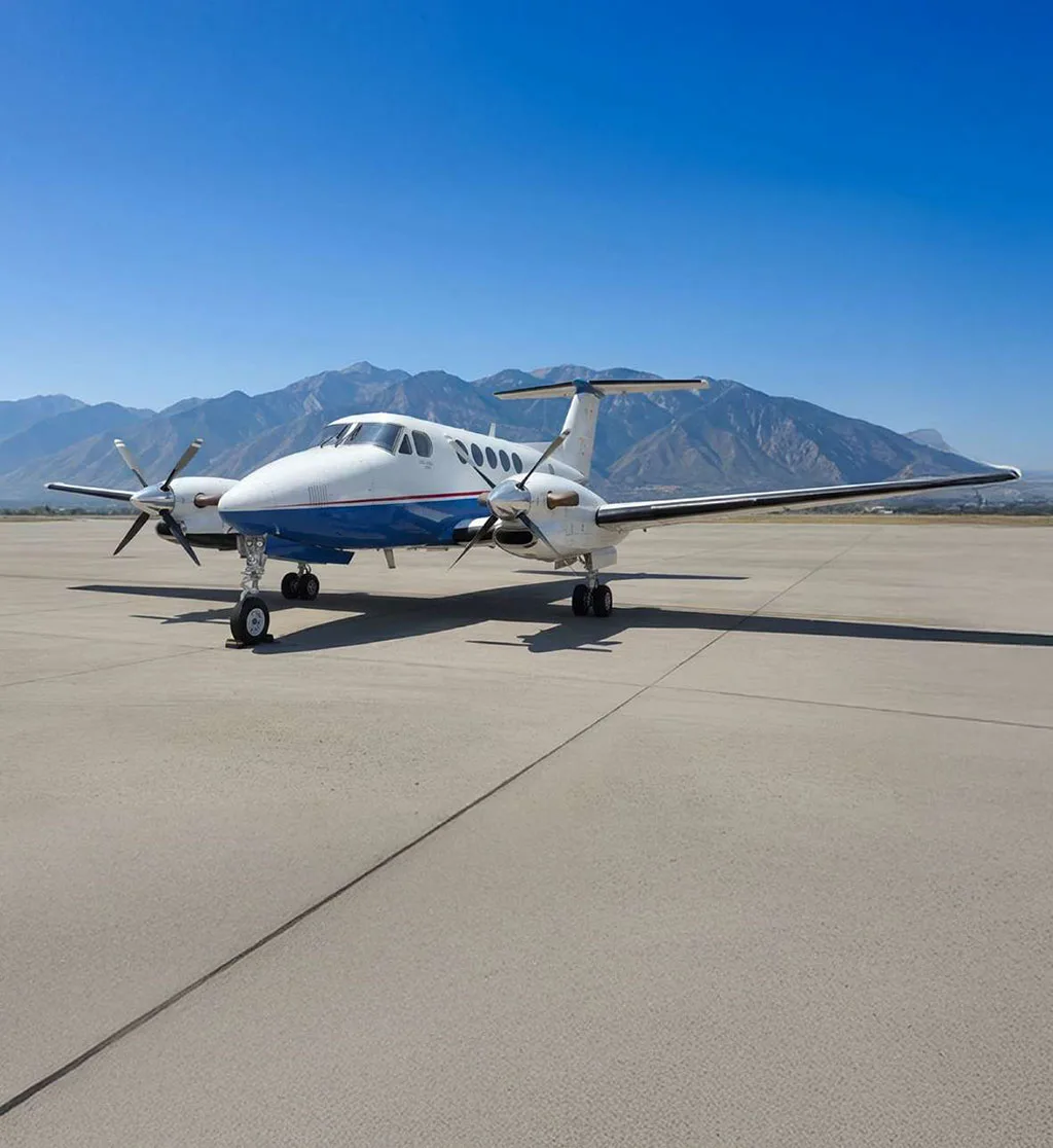 A white and blue turboprop aircraft, likely a King Air, seen from the front on an asphalt runway with mountains in the background.
