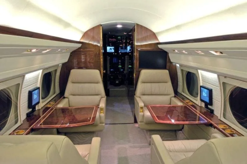 A view down the aisle of a private jet cabin with two beige leather seats facing each other across a wooden table, with the cockpit visible in the background.