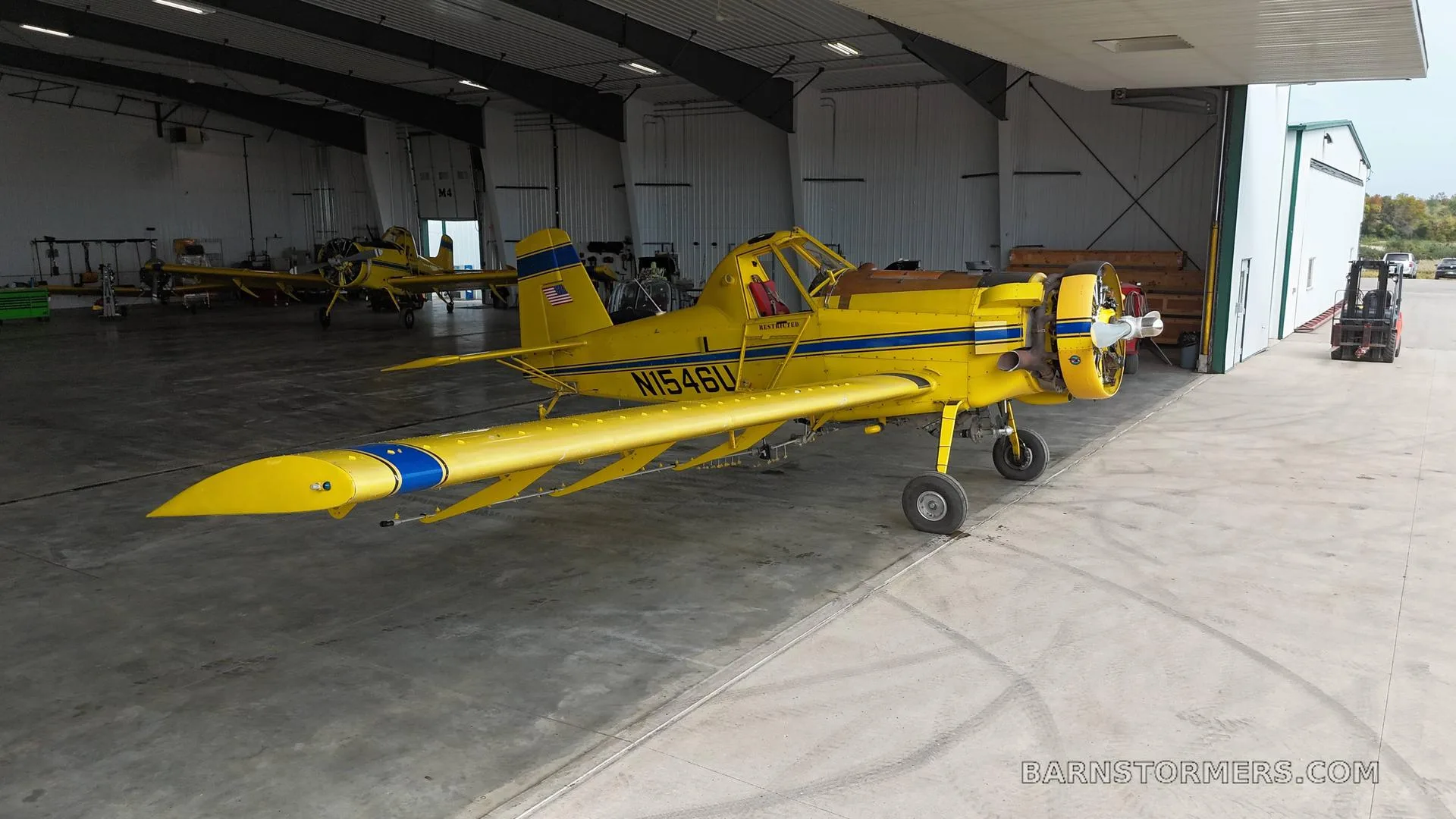 A yellow agricultural aircraft with blue stripes (N1546U) parked inside a large hangar with the door open, showing other aircraft in the background.