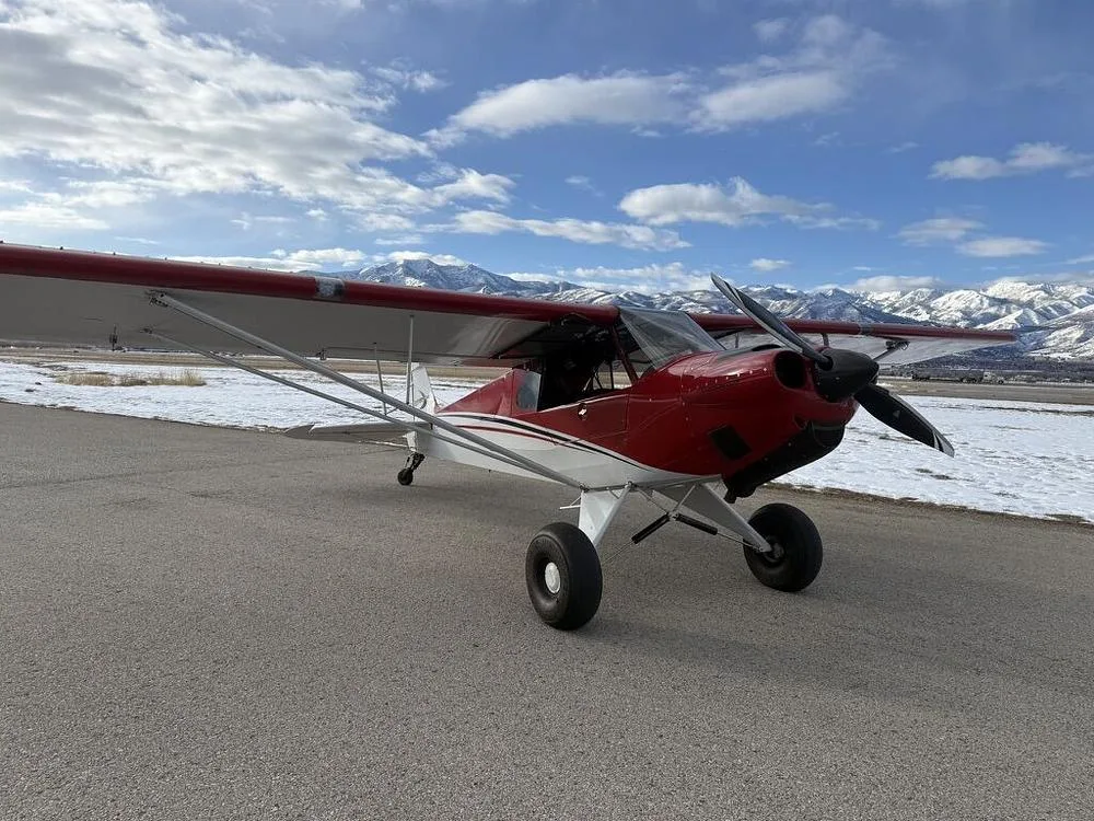 A red and white bush plane with large tires parked on an asphalt runway with snow-capped mountains in the background.