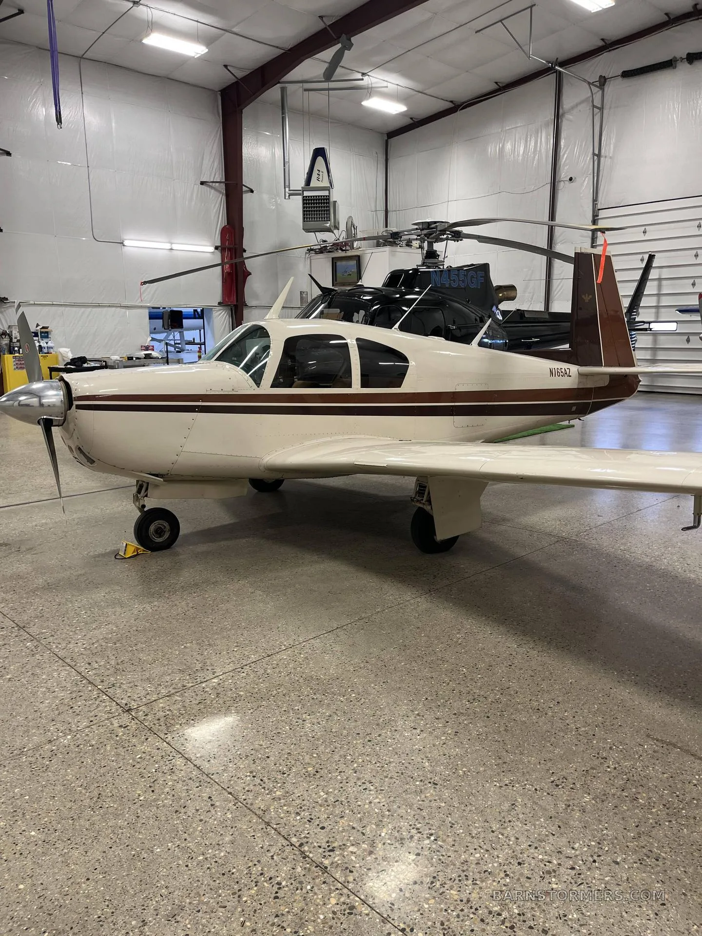 A cream and brown single-engine propeller aircraft (Beechcraft Debonair N165AZ) is parked in a hangar, with a black helicopter in the background.