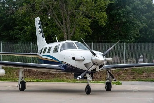 A white and blue single-engine turboprop aircraft is parked on a tarmac with trees and a fence in the background.