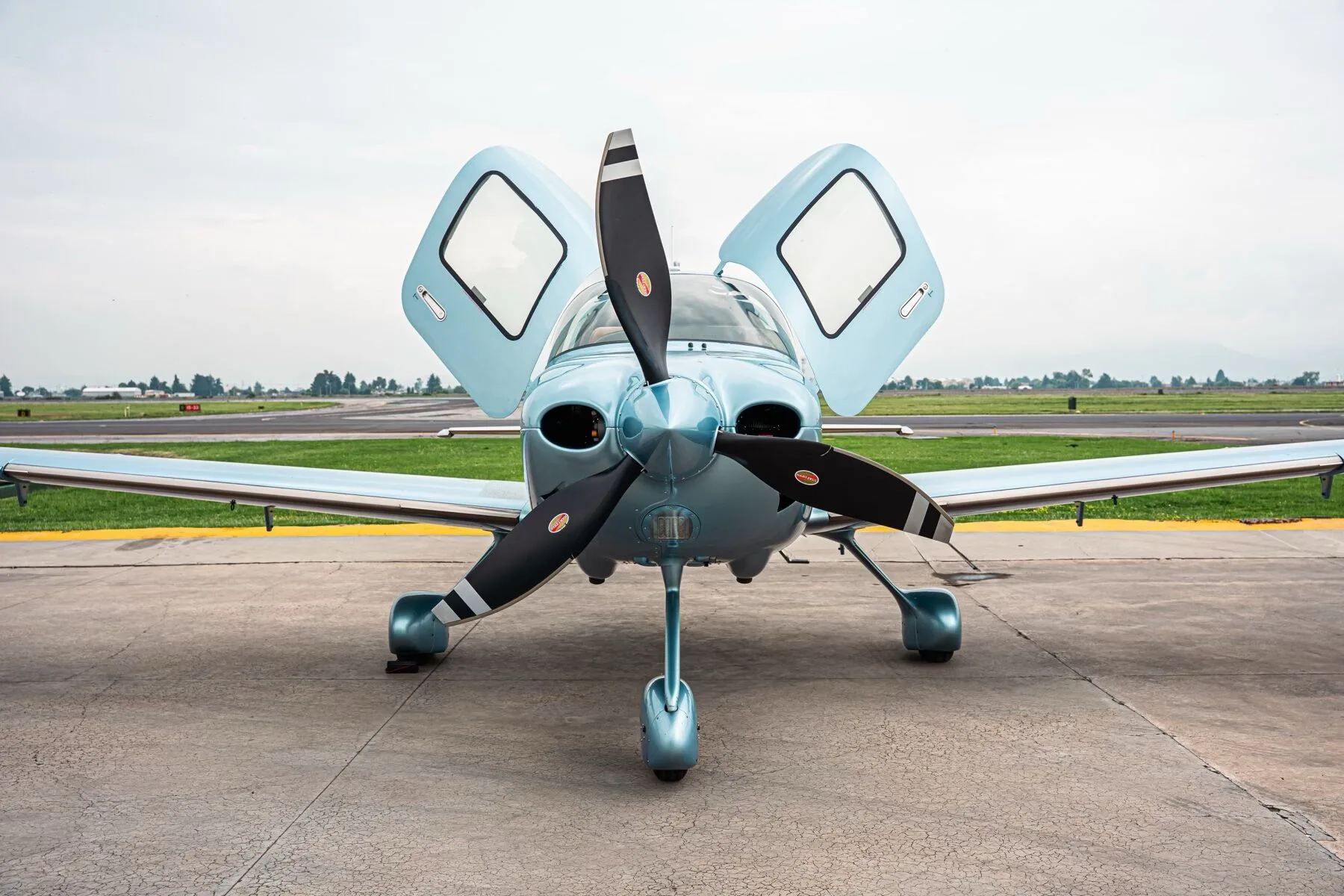 Front view of a light blue Cirrus SR22T aircraft with its gull-wing doors open on an airfield.