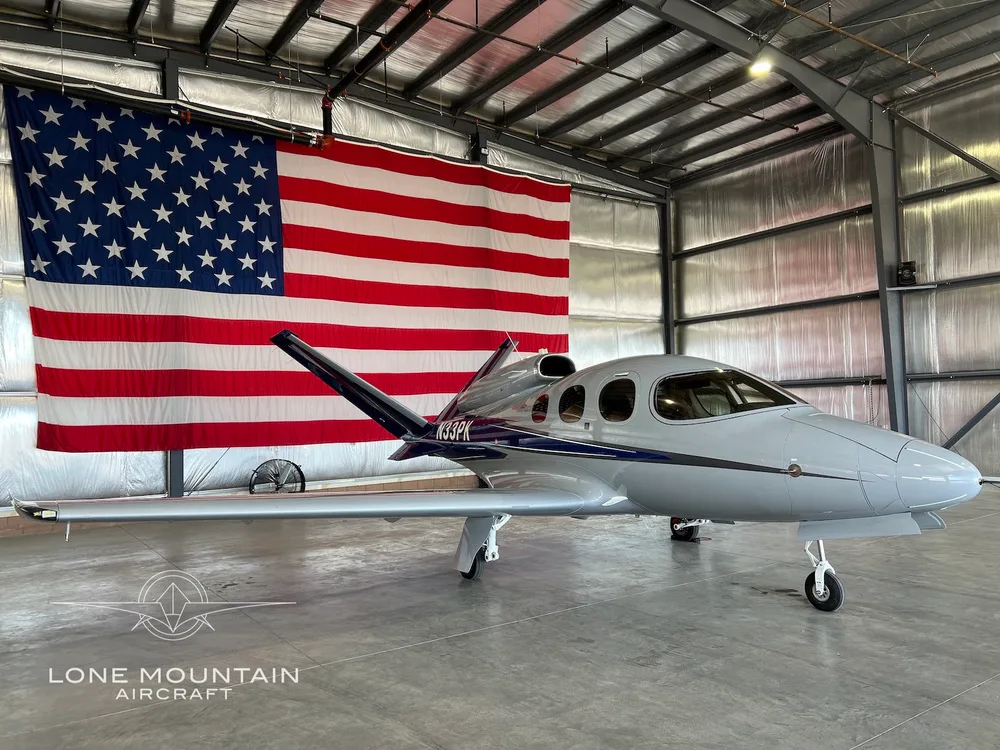 A grey and blue Cirrus Vision Jet (N33PK) is parked in a hangar in front of a large American flag.