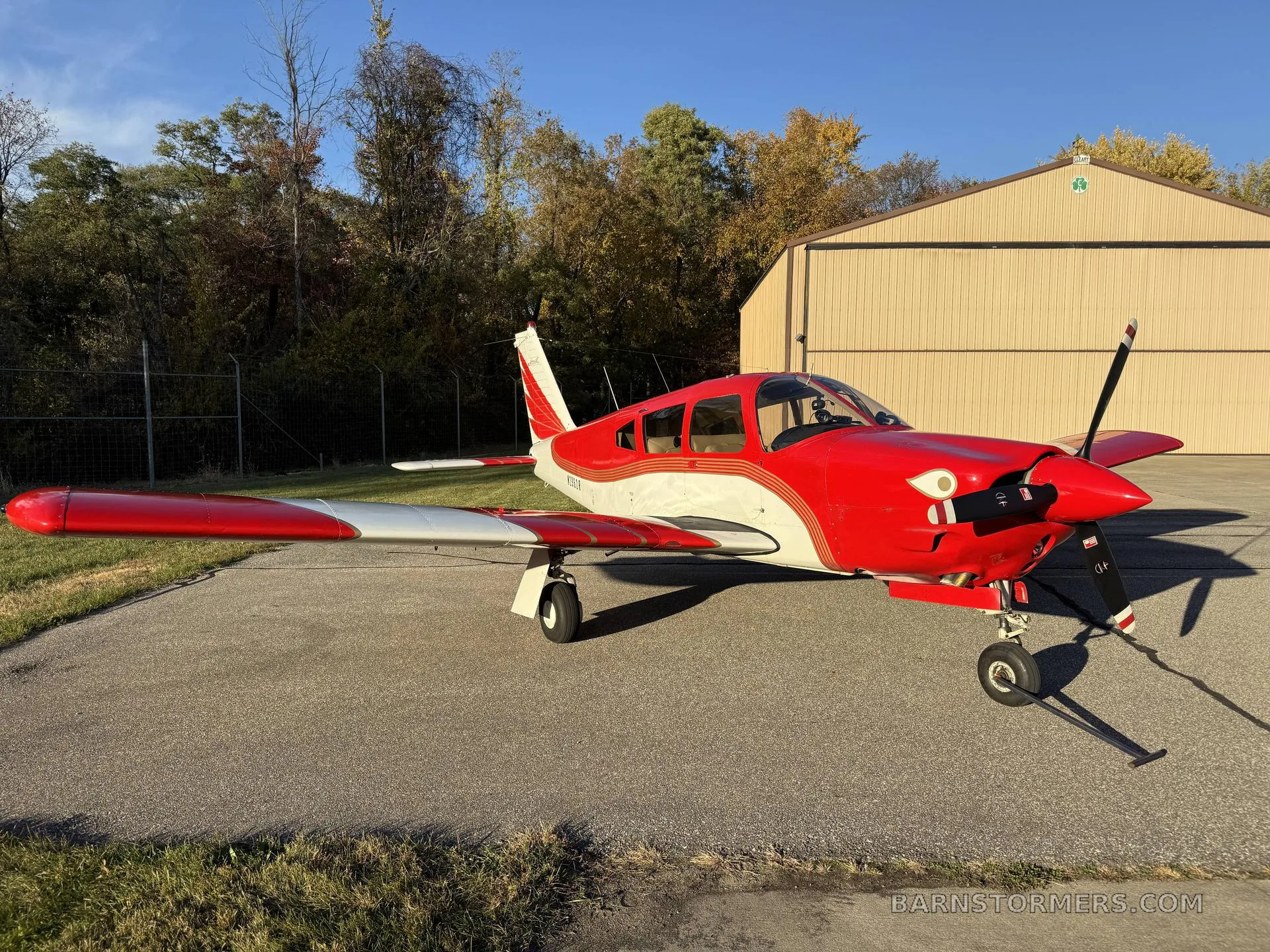 A red and white Mooney M20 series aircraft parked on an asphalt apron outside a hangar.